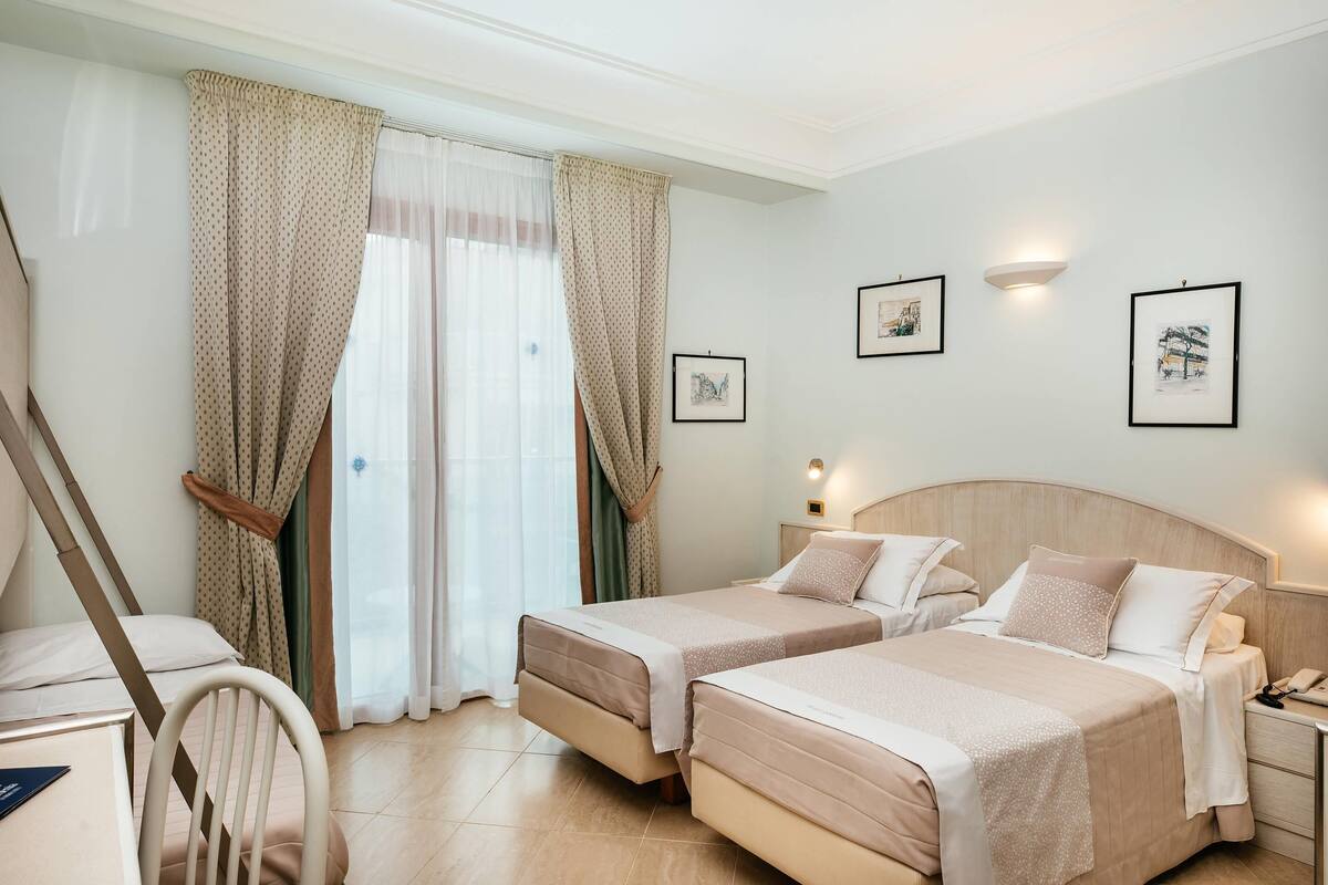 Une chambre d’hôtel avec deux lits, un bureau, une chaise et une fenêtre avec des rideaux. Les murs sont ornés de tableaux encadrés, et il y a une table de chevet avec une lampe.