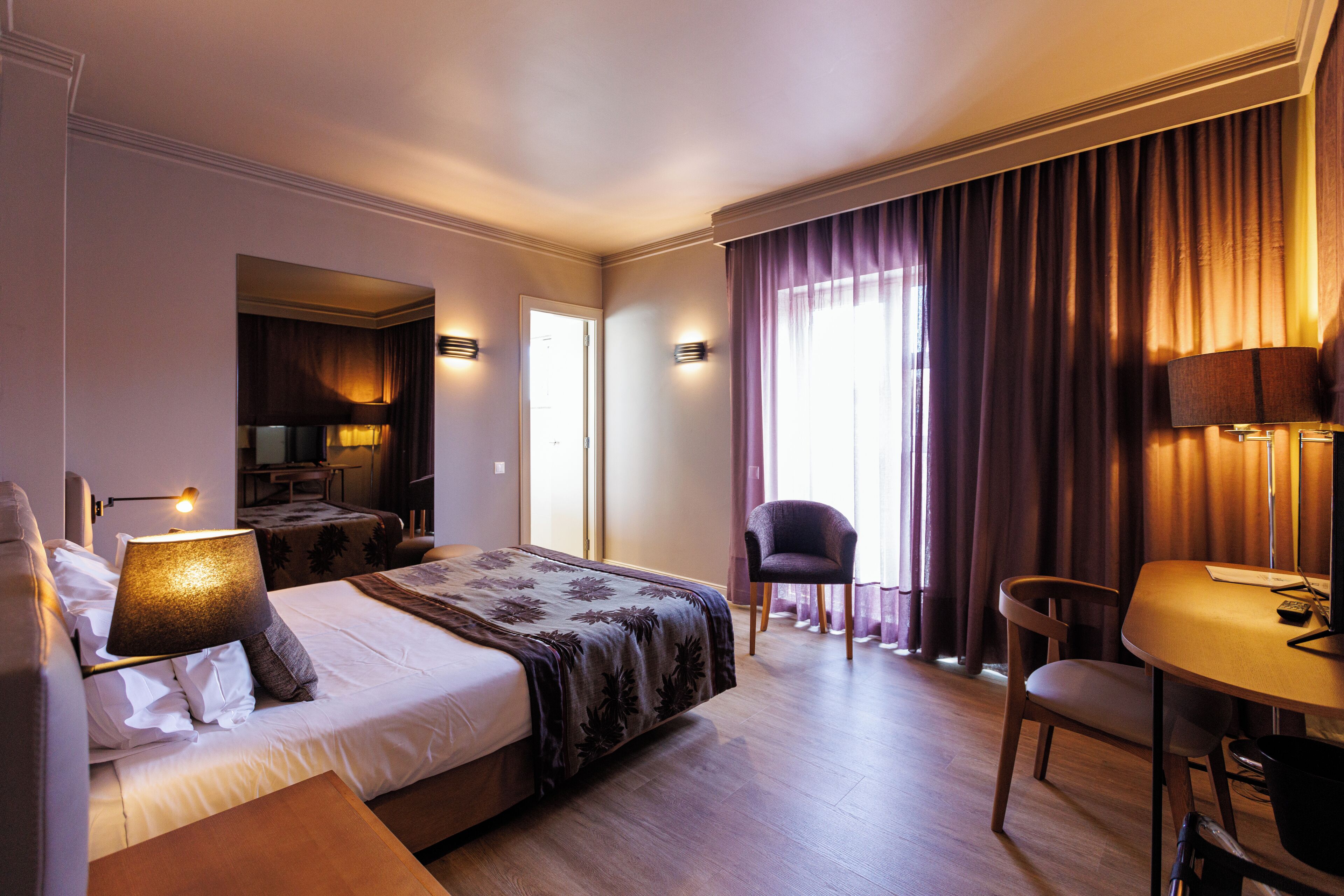 A hotel room with a bed, a desk, a chair, and a lamp.