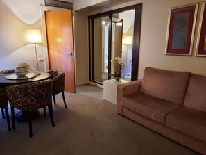A hotel room with a sofa, a dining table with chairs, a small table with a vase, and a door leading to a bathroom.