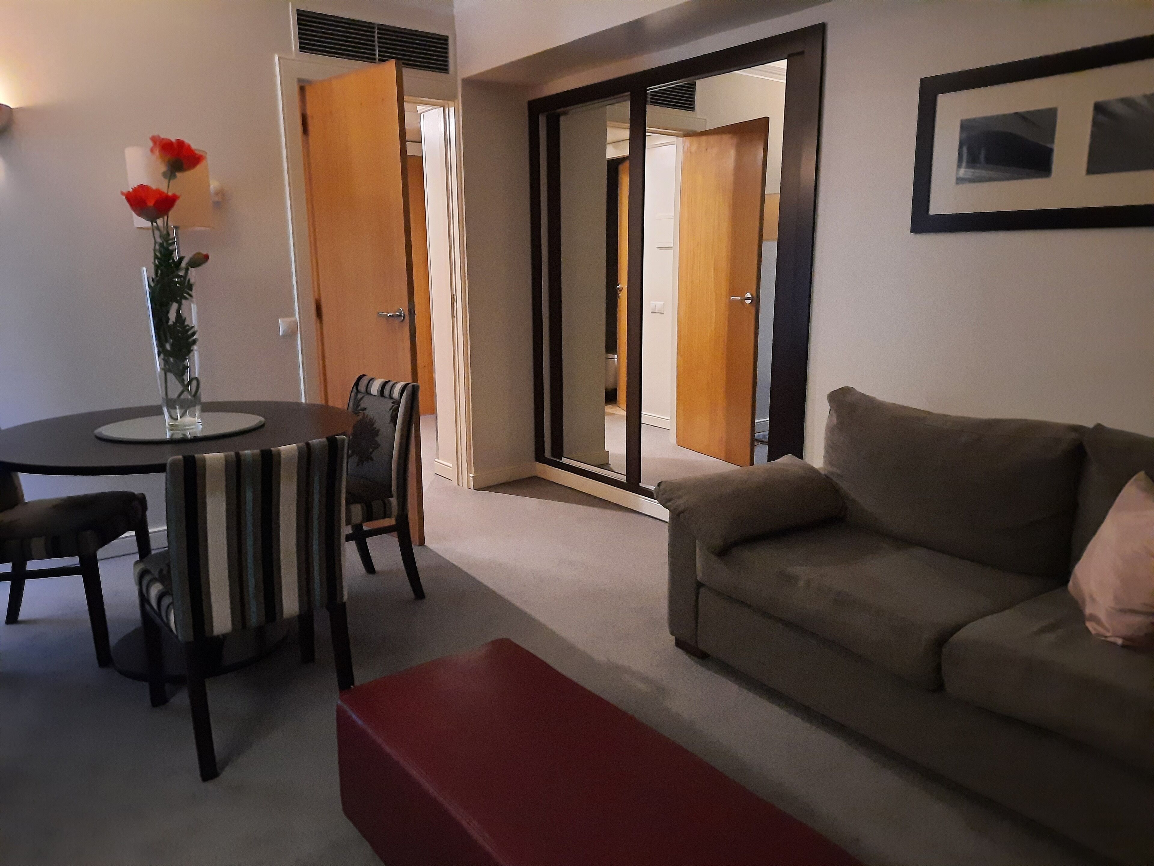 A living room with a sofa, a dining table with chairs, and a red bench. There is a vase with flowers on the table.