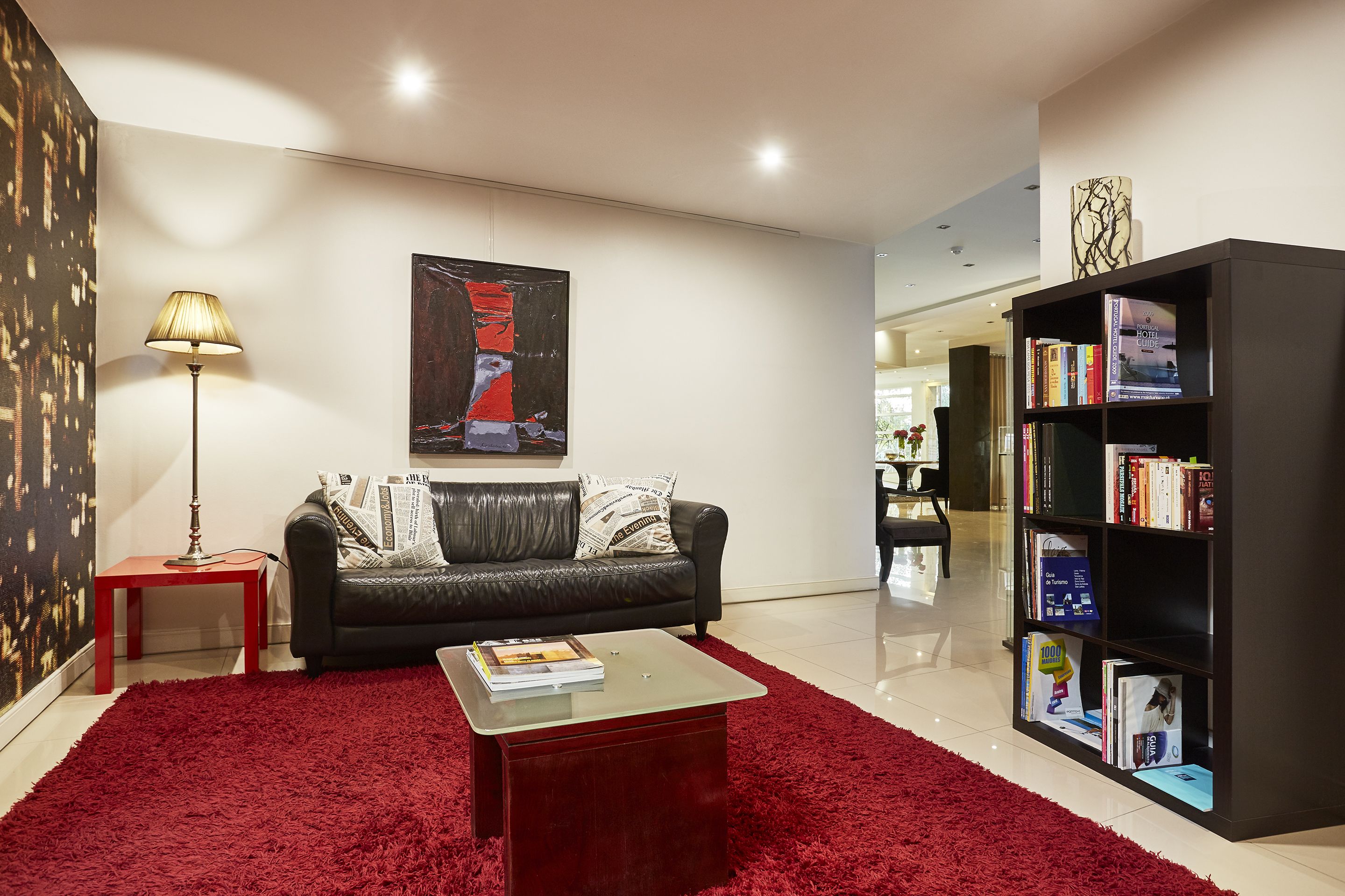 Una sala de estar moderna con un tapete de pelo rojo, un sofá de cuero negro, una mesa de centro de vidrio, una lámpara de pie, un cuadro abstracto grande y una estantería con libros y una guía hotelera.