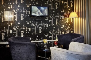 A modern interior with a TV mounted on a textured wall, a lamp, and two chairs around a small table with drinks.