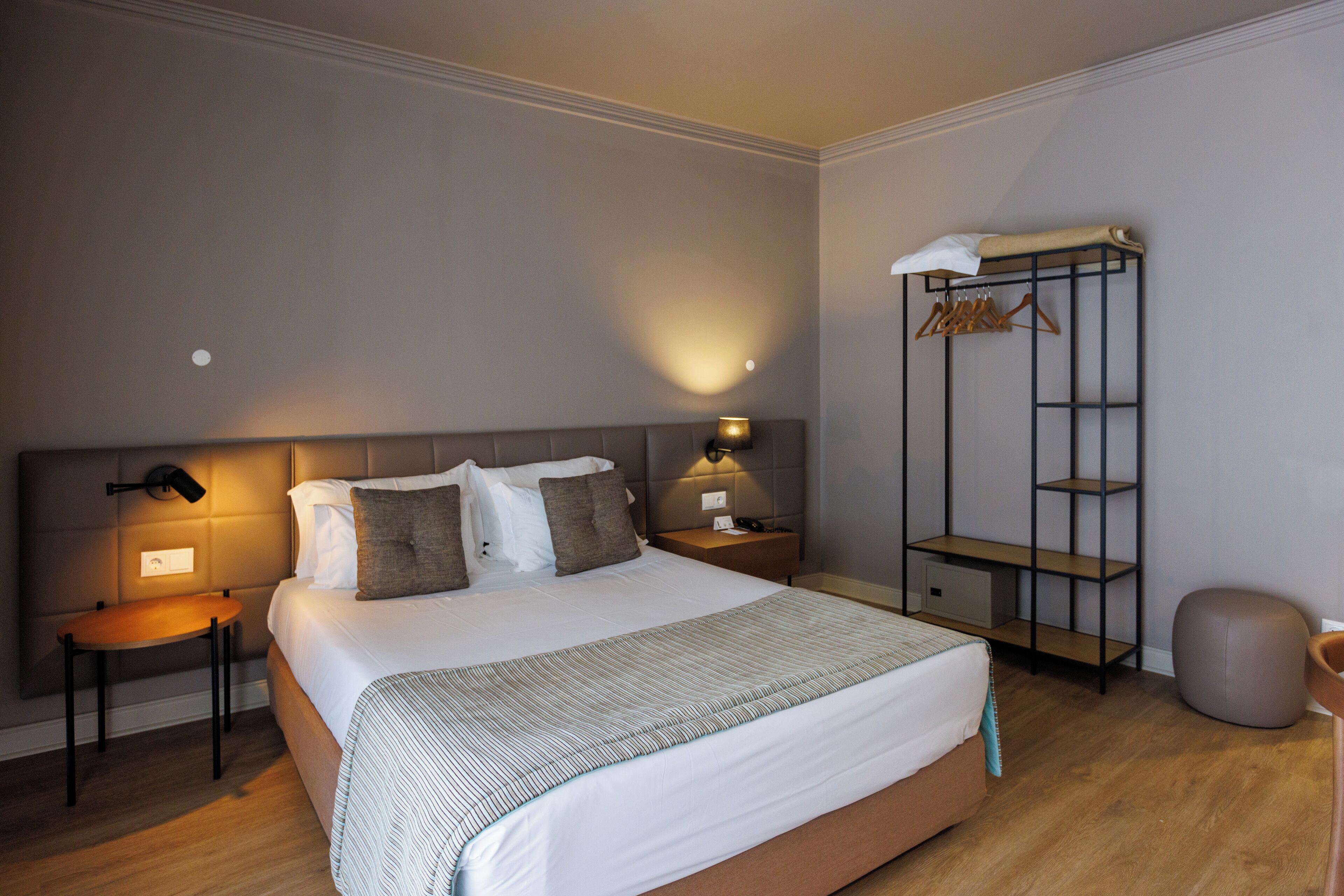 Quarto de hotel bem arrumado, com uma cama grande, mesas de cabeceira com abajures, um guarda-roupa e um pequeno banquinho.