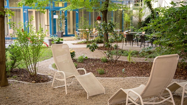A patio area with two beige lounge chairs, a small garden with plants and a tree, and a building with blue doors and windows in the background. - AllYouNeed Hotel Vienna 2 (Vienna)