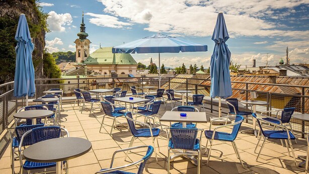 An outdoor terrace with blue chairs and tables, a view of a church with a green roof, and a clear sky. - Allyouneed Hotel Salzburg (Salzburg)