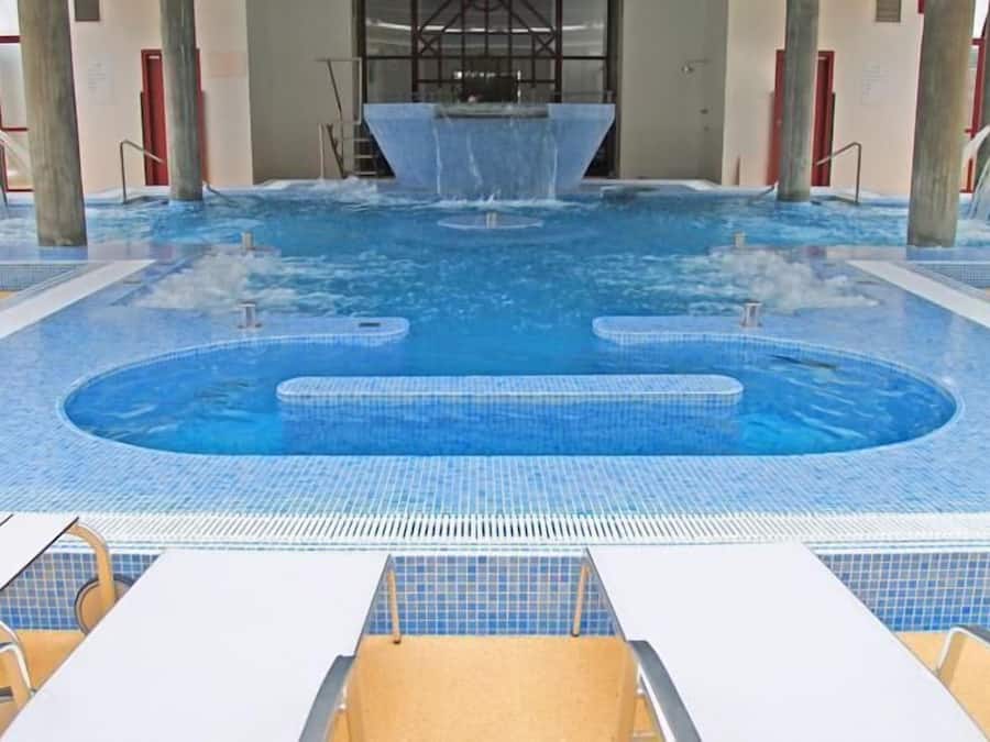 A large indoor pool with a central water feature, multiple entry points, and lounge areas with chairs.