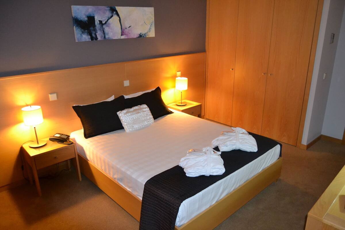 A neatly made bed with white linens, two bedside lamps, a telephone, and a folded towel on the bed.