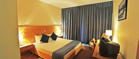 Habitación de hotel con cama, escritorio, silla, televisión y ventana con cortinas.