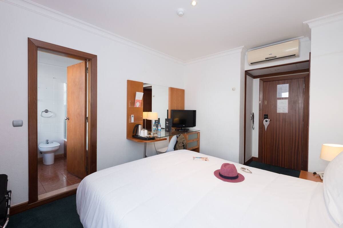 Ein Hotelzimmer mit einem Bett, einem Fernseher, einem Schreibtisch mit Lampe, einem Spiegel und einer Tür zu einem Badezimmer.