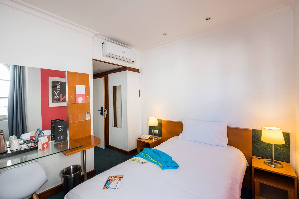 Ein Hotelzimmer mit einem Bett, Nachttischen, einem Schreibtisch, einem Sessel, einer Mülltonne und einer Tür zu einem anderen Raum.
