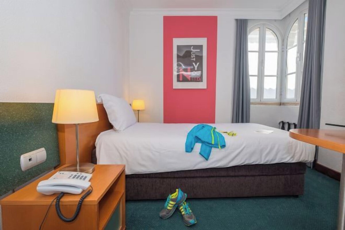 A hotel room with a bed, a bedside table with a lamp, a phone, a pair of shoes, and a window with curtains.
