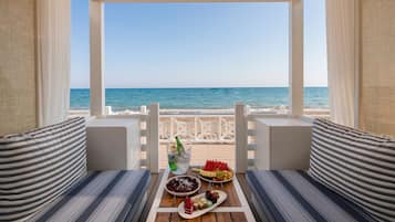 Ein Balkon direkt am Strand mit Meerblick, ein Esstisch mit Snacks und einer Flasche sowie gestreifte Kissen.