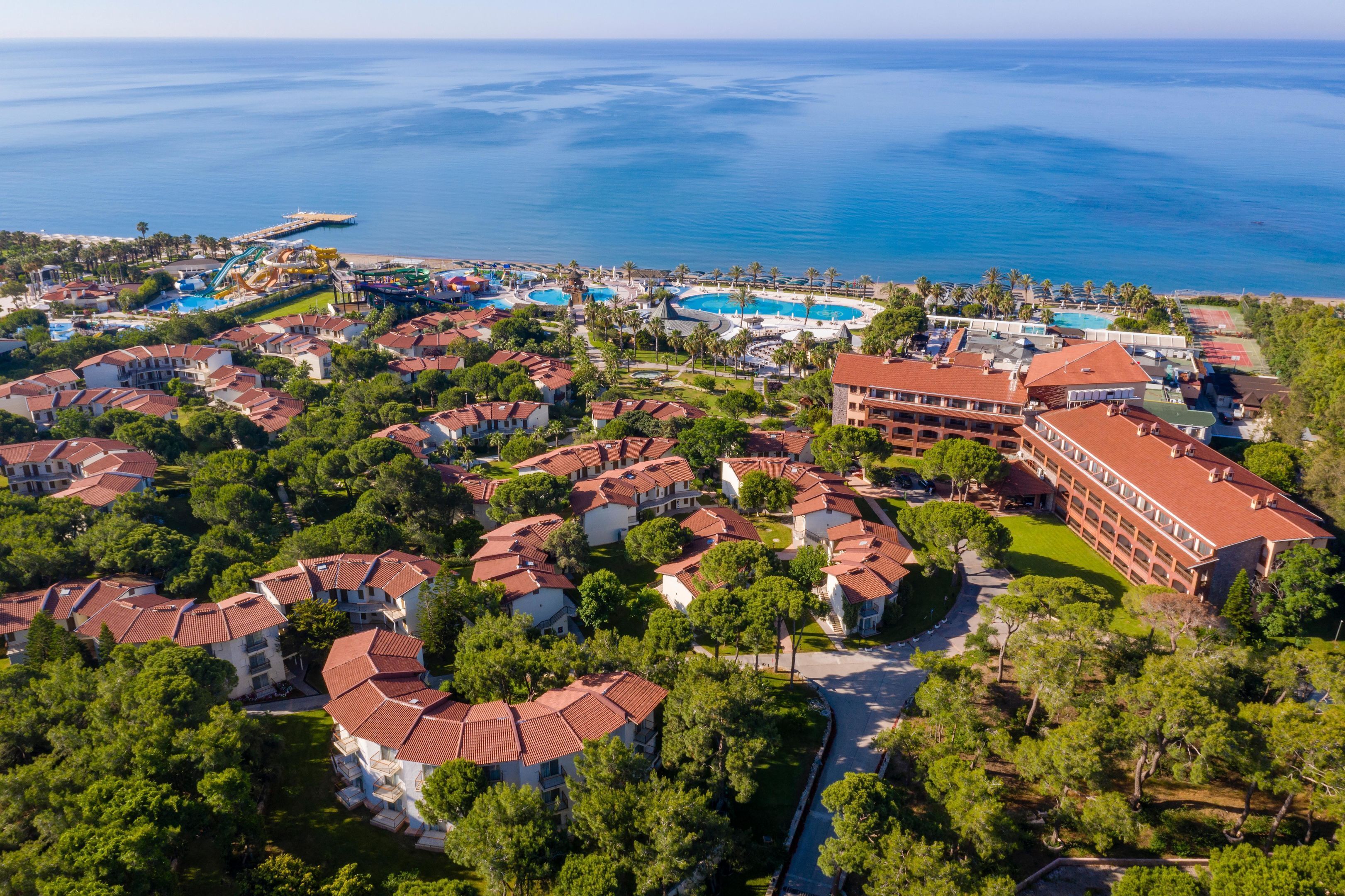 Vista aerea di un resort costiero con diversi edifici, un ampio complesso di piscine e un molo che si protende verso il mare.