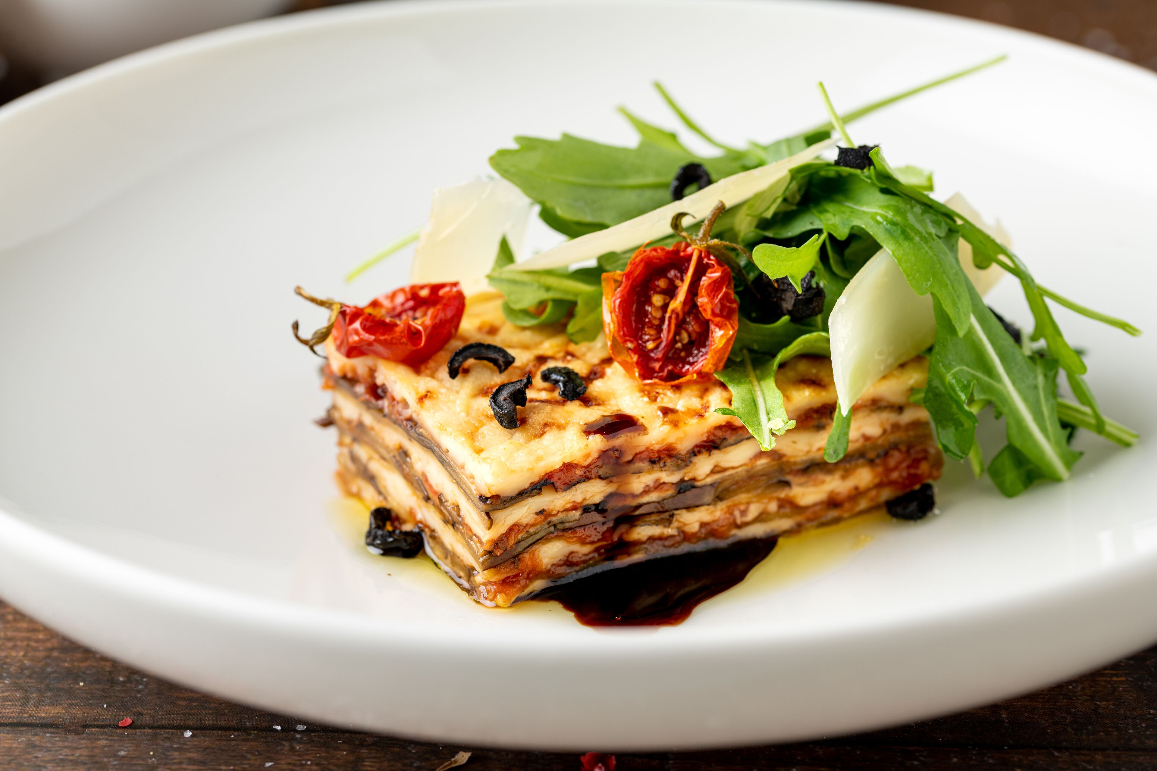 A slice of lasagna on a white plate with cherry tomatoes, black olives, and arugula.