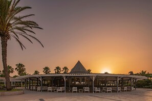 A covered outdoor seating area with benches and tables, surrounded by palm trees and a sunset in the background. - Papillon Belvil Holiday Village - All Inclusive (Serik)