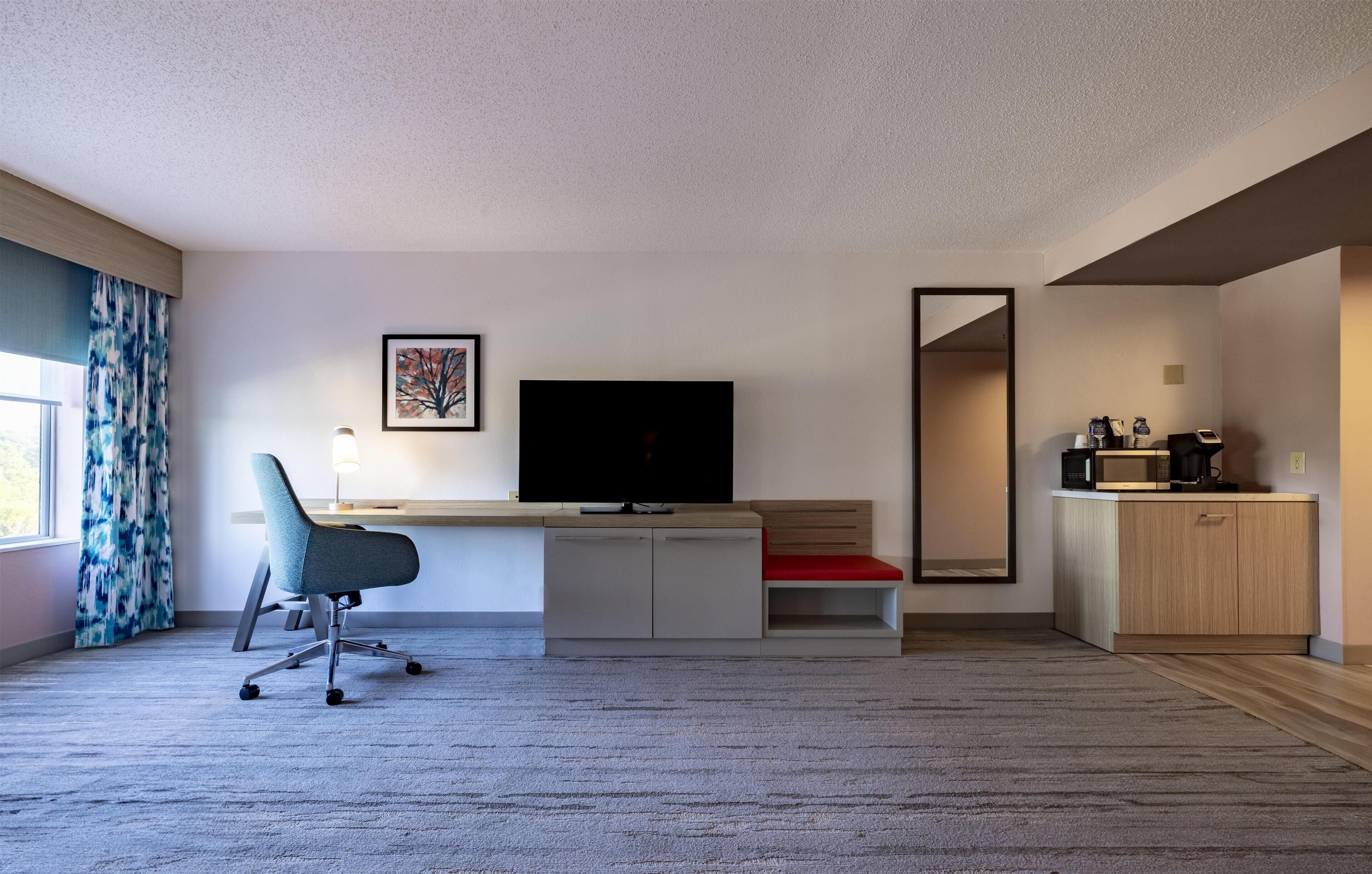 Quarto de hotel moderno com uma televisão de tela plana grande, uma escrivaninha com cadeira, um banco acolchoado vermelho, uma cozinha compacta com micro-ondas e um espelho grande.