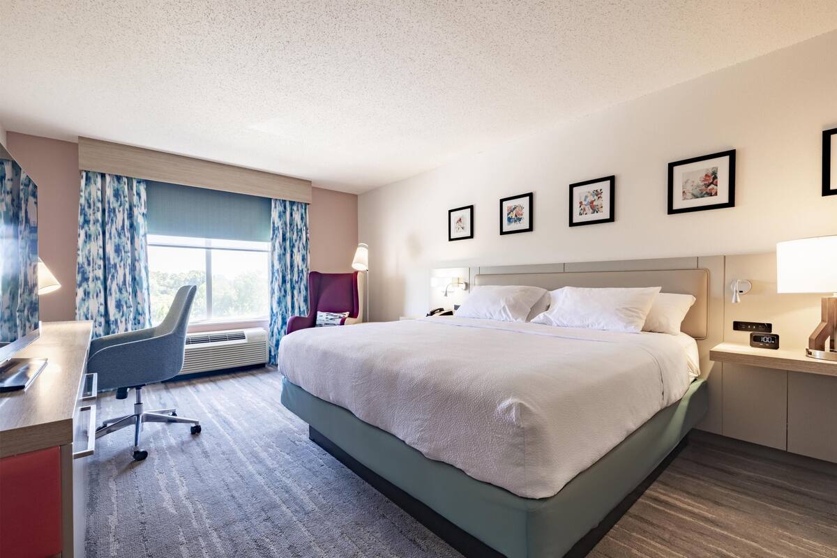 A hotel room with a large bed, a desk, a chair, and a window with curtains.