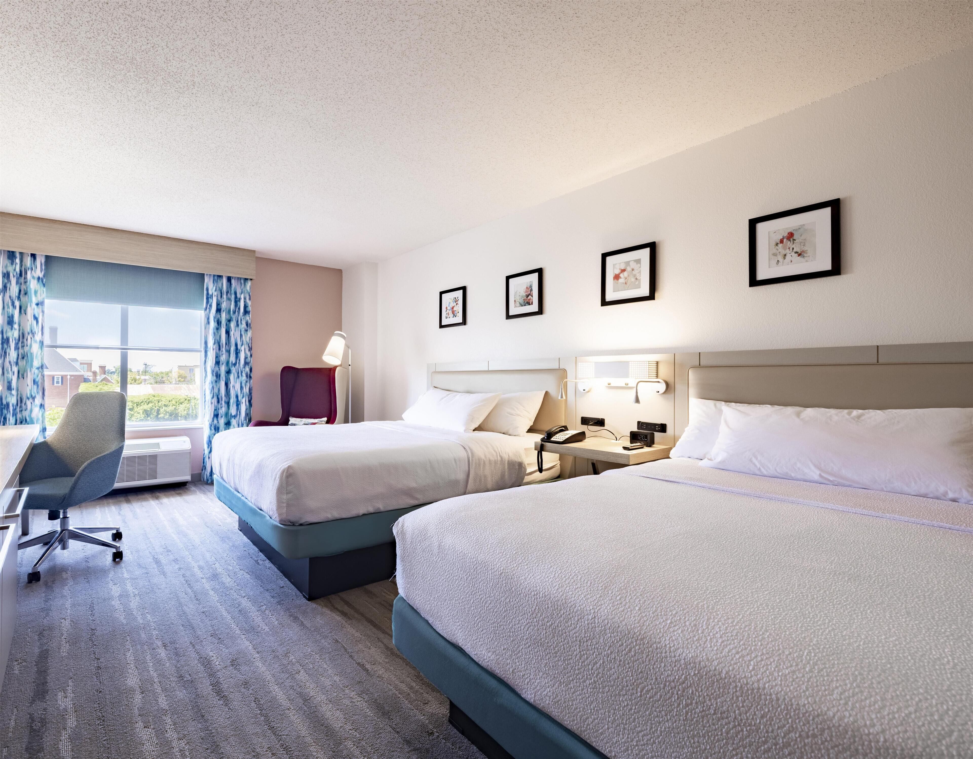 Habitación de hotel con dos camas, un escritorio con lámpara, una silla y una ventana con cortinas.