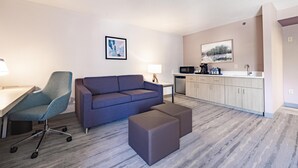 A modern hotel room with a sofa, a desk, a chair, and a kitchenette.