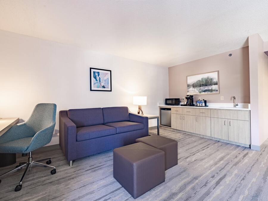 A modern hotel room with a sofa, a desk, a chair, and a kitchenette.