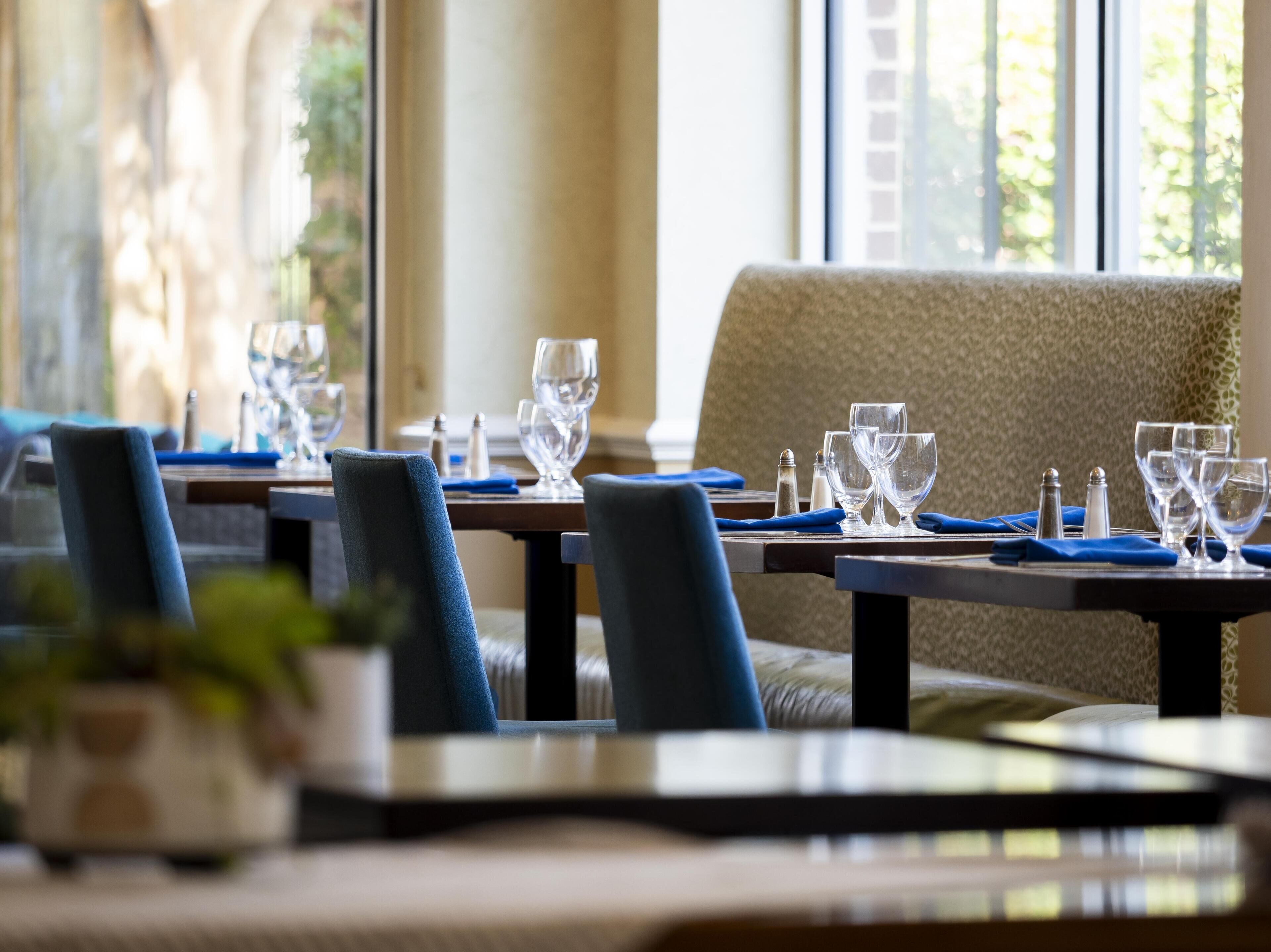 A dining area with tables set for two, featuring blue upholstered chairs and clear glassware.