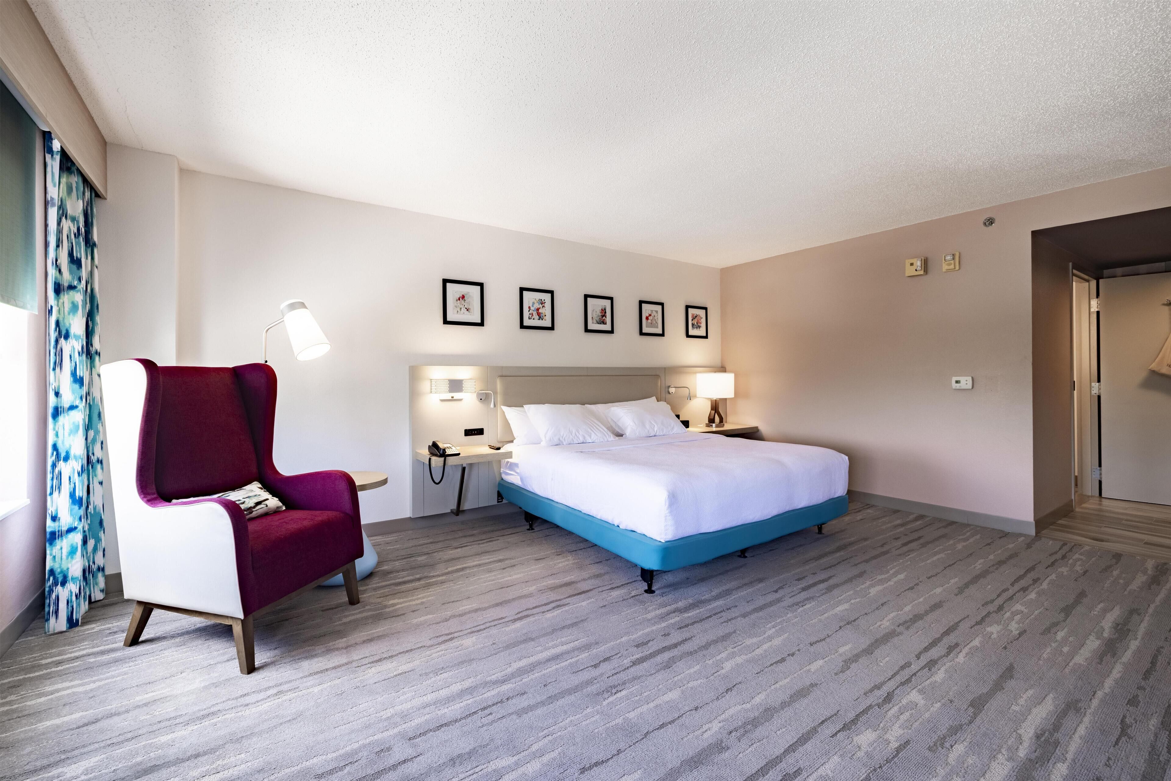 A hotel room with a bed, a red armchair, a desk, and a nightstand.