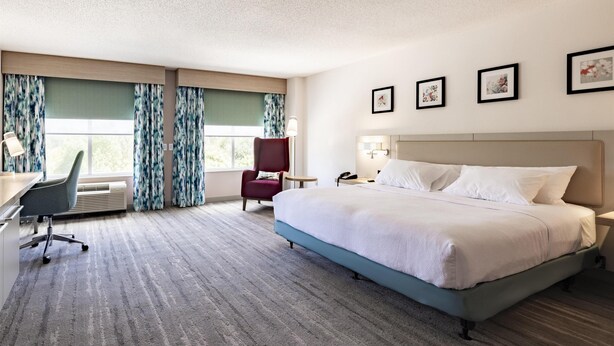 Quarto de hotel com uma cama grande, uma mesa com cadeira, uma luminária de mesa e uma janela com cortinas. Há três quadros na parede.