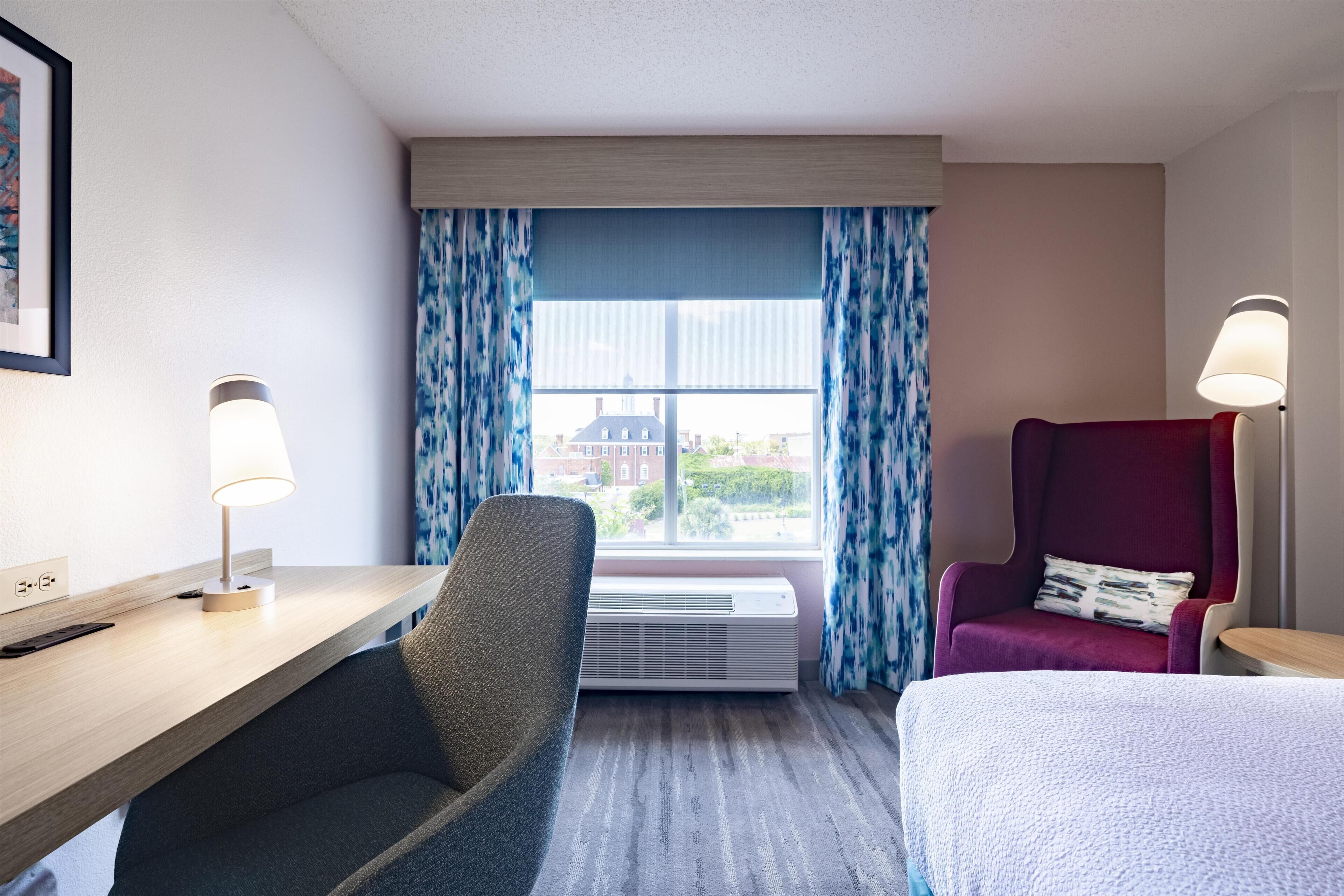 A hotel room with a desk, a chair, a bed, a window with curtains, and a lamp.