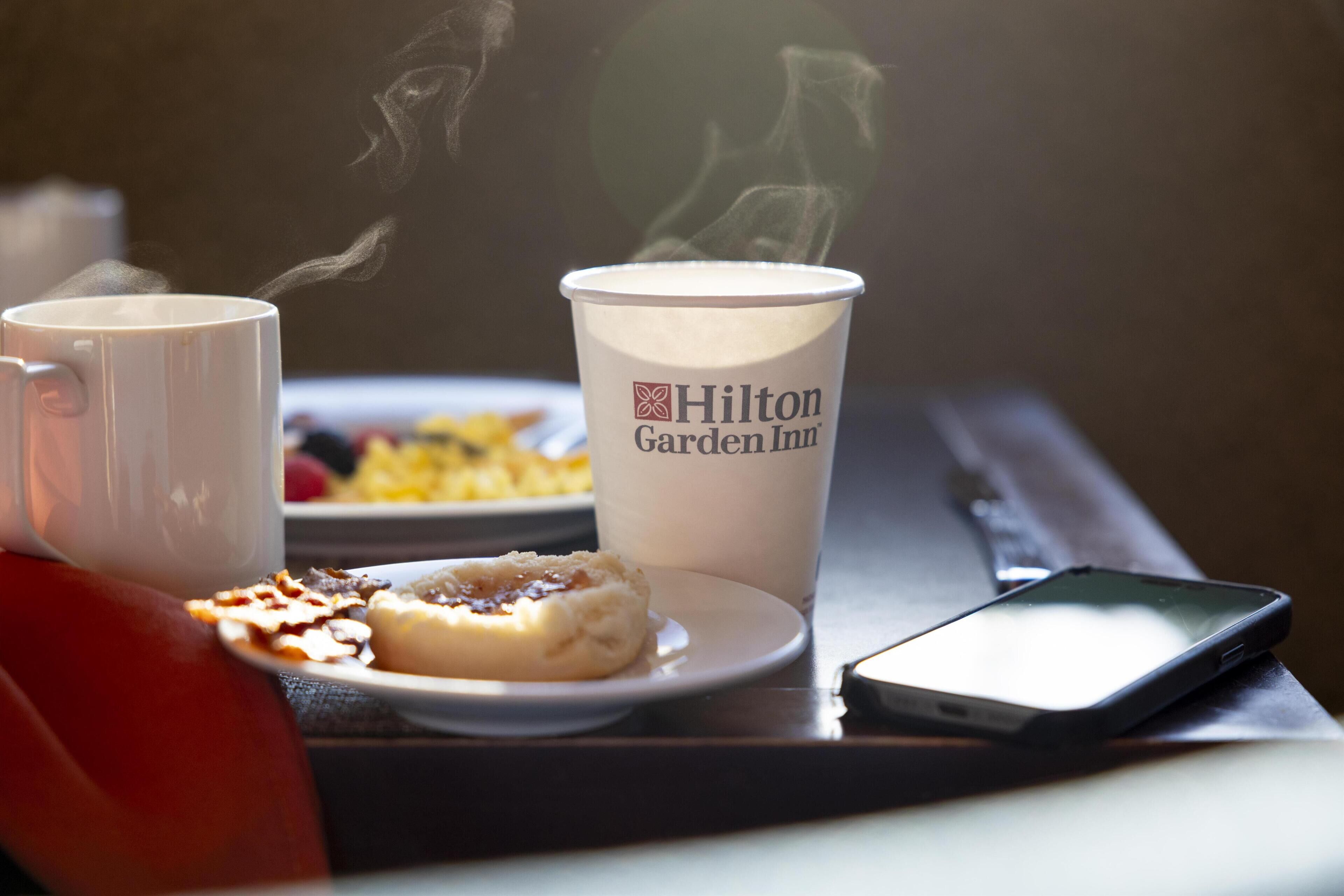 Una mesa con una taza de Hilton Garden Inn, un pastel y un teléfono inteligente.