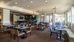 A hotel dining area with multiple tables and chairs, a kitchen in the background, and large windows allowing natural light.