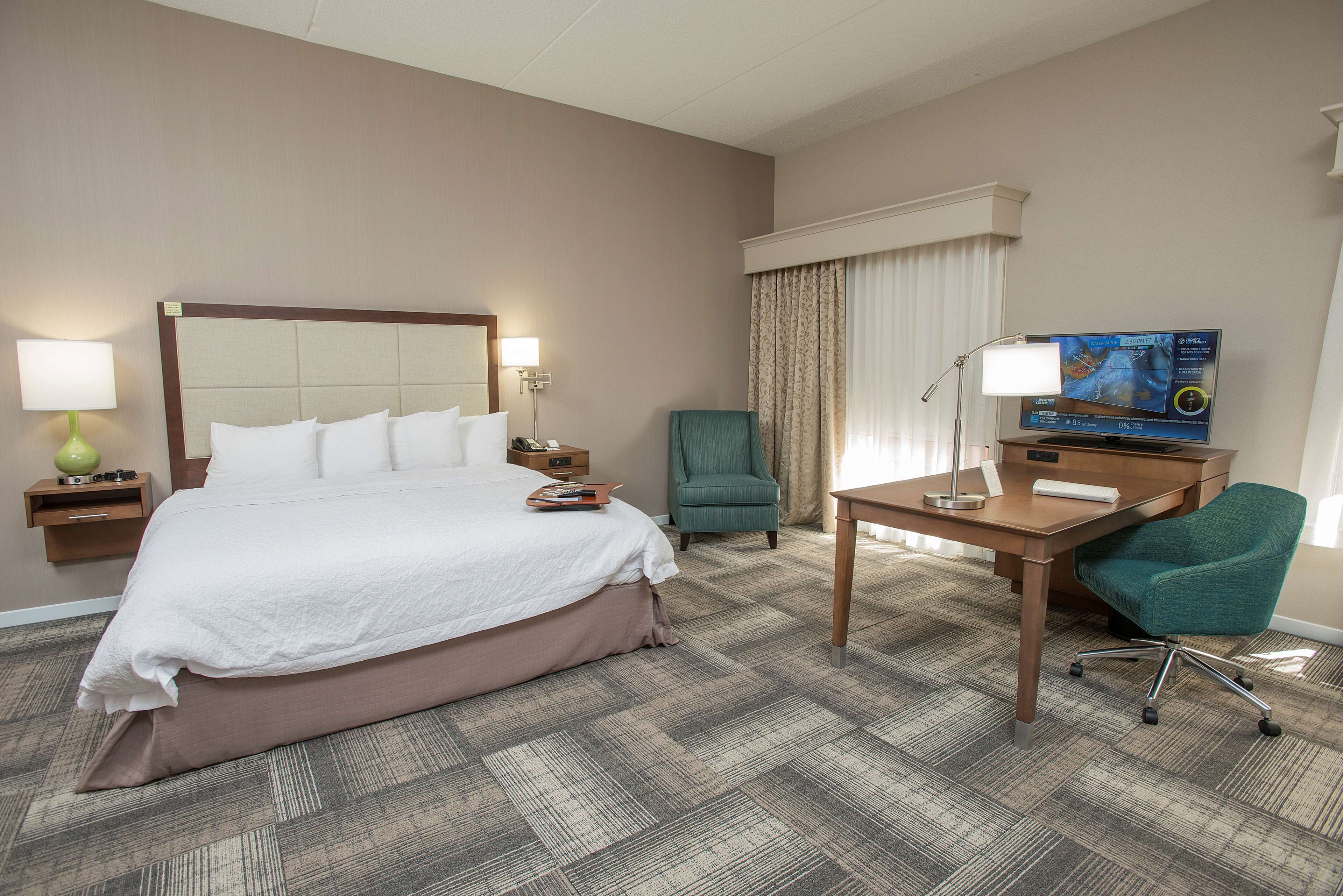 A hotel room with a large bed, a desk, a chair, a TV, and a lamp.