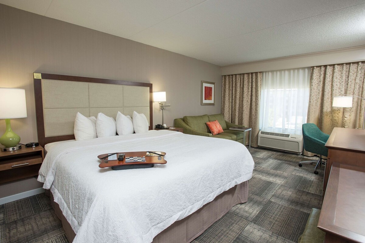 Habitación de hotel con una cama grande, una mesita de noche con lámpara, un sillón verde, un escritorio con silla y una ventana con cortinas.