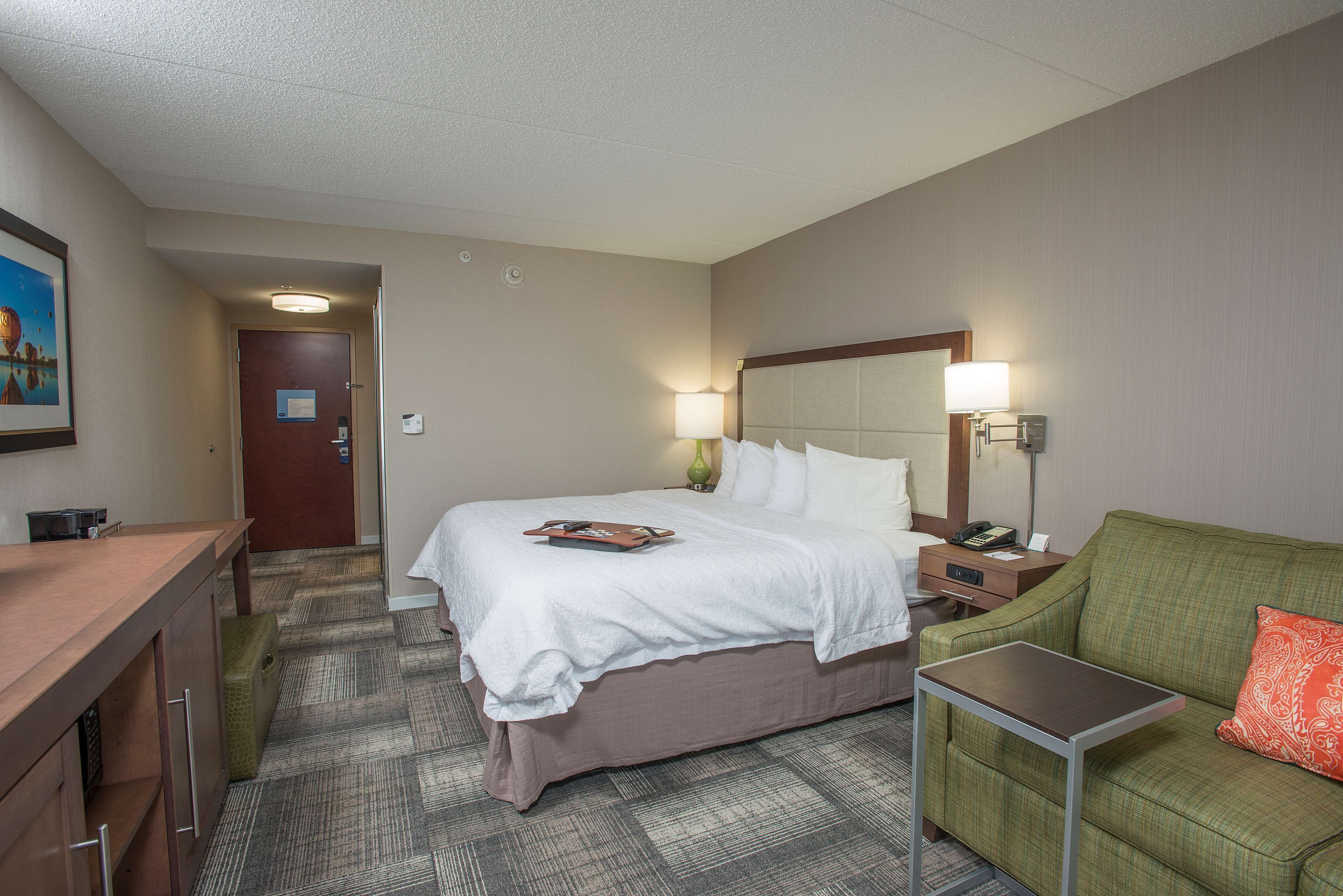 A hotel room with a bed, bedside table, lamp, and a green sofa with an orange pillow.