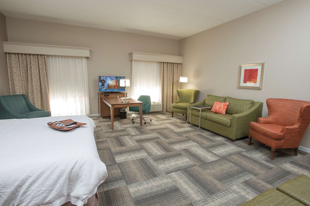 A hotel room with a bed, a desk, a television, a green armchair, a red armchair, and a green sofa.