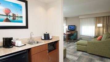 Habitación de hotel con kitchenette, televisor, sofá verde y un cuadro con globos aerostáticos.
