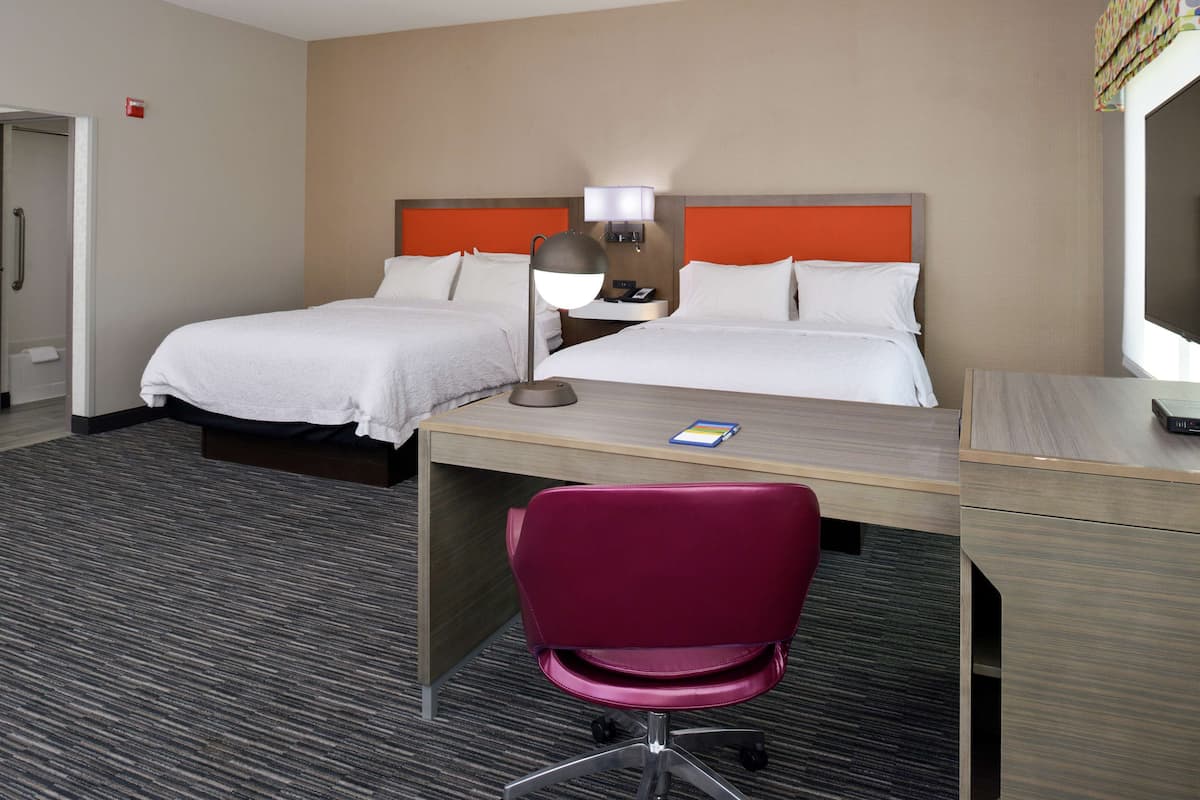 A hotel room with two beds, a desk, a chair, a lamp, and a television.