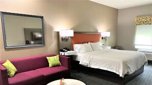 A hotel room with a bed, a red sofa with a green pillow, a round table, and a large mirror. - Hampton Inn Fort Stockton (Fort Stockton)