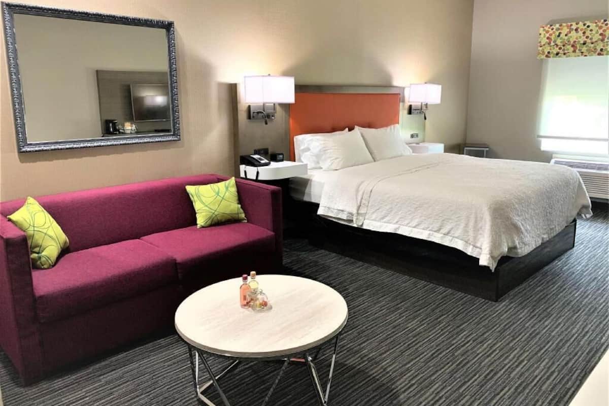 A hotel room with a bed, a sofa, a round table, a nightstand, a lamp, and a mirror.