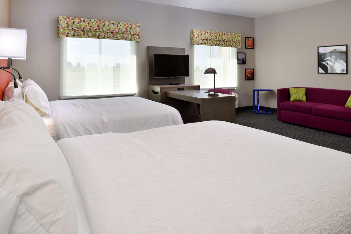 A hotel room with two beds, a desk with a flat-screen TV, a red sofa, and large windows with colorful valances.