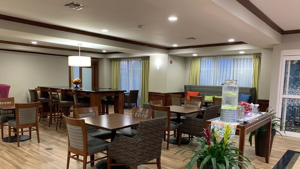 A hotel dining area with wooden tables and chairs, a large potted plant, and a view of the outdoors through a window. - Hampton Inn Fort Stockton (Fort Stockton)