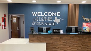 A reception desk with a 'Welcome to the Lone Star State' sign, HP and Hilton Honors logos, a small bird decoration, and a fire extinguisher on the wall. - Hampton Inn Fort Stockton (Fort Stockton)
