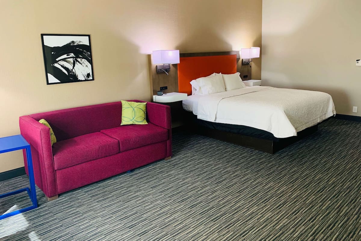 A hotel room with a bed, a red sofa, a small table, and a wall-mounted picture.
