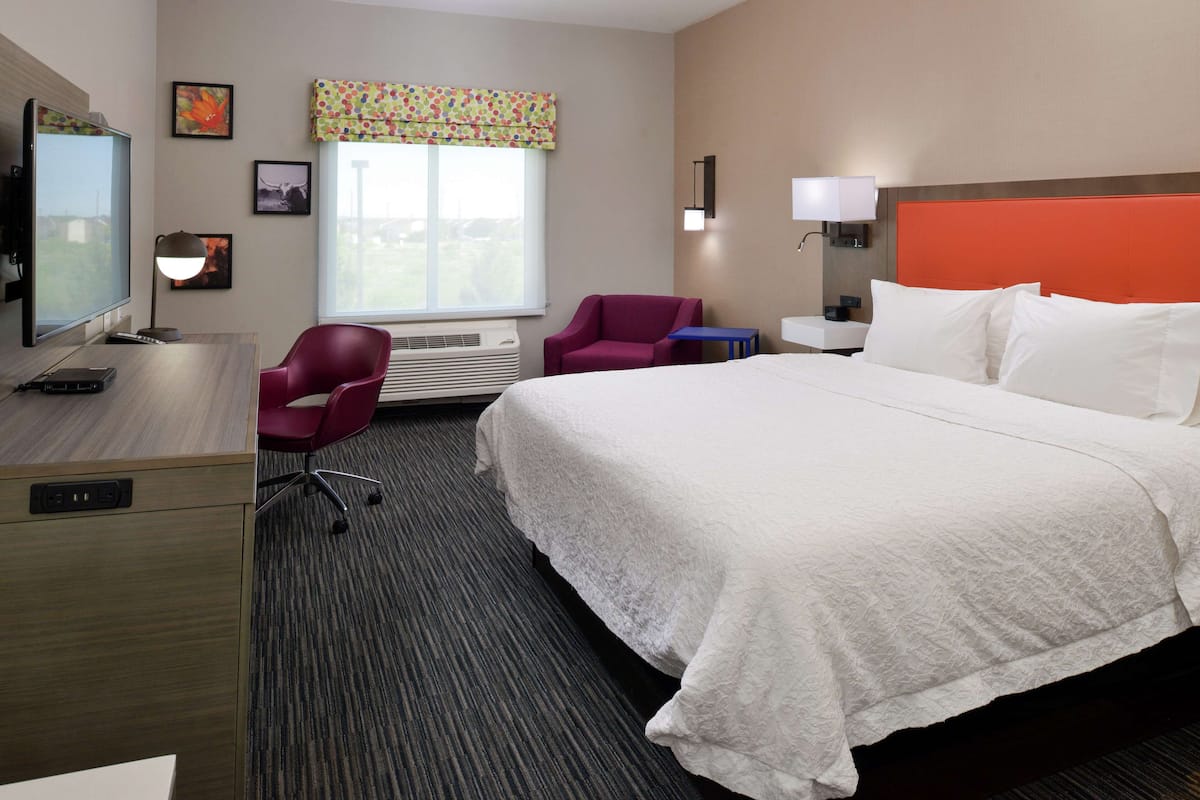 A hotel room with a large bed, a desk with a TV, a chair, a window with curtains, and a nightstand with a lamp.