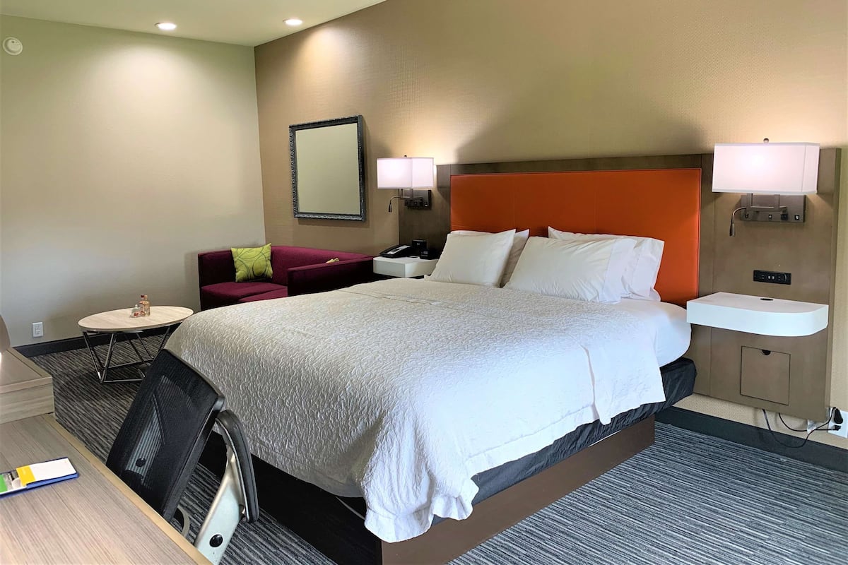 A hotel room with a large bed, a desk, a chair, a small table, a sofa, a mirror, and a nightstand with a lamp.