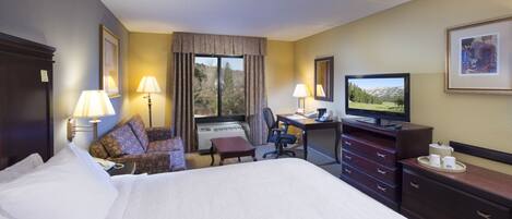 Una habitación de hotel con una cama grande, un escritorio con computadora, una silla, televisor y ventana con cortinas.