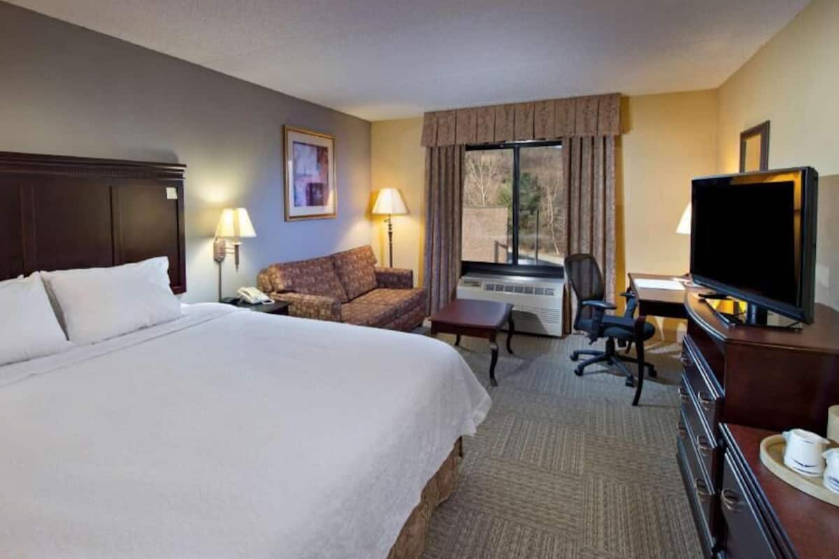A hotel room with a large bed, a sofa, a desk with a computer, a TV, and a window with curtains.