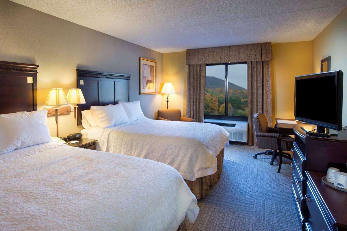 A hotel room with two beds, a desk with a television, and a window with a mountain view.