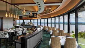 En restaurant med buet indretning, store vinduer med udsigt over vand, og et barområde med udsigt.