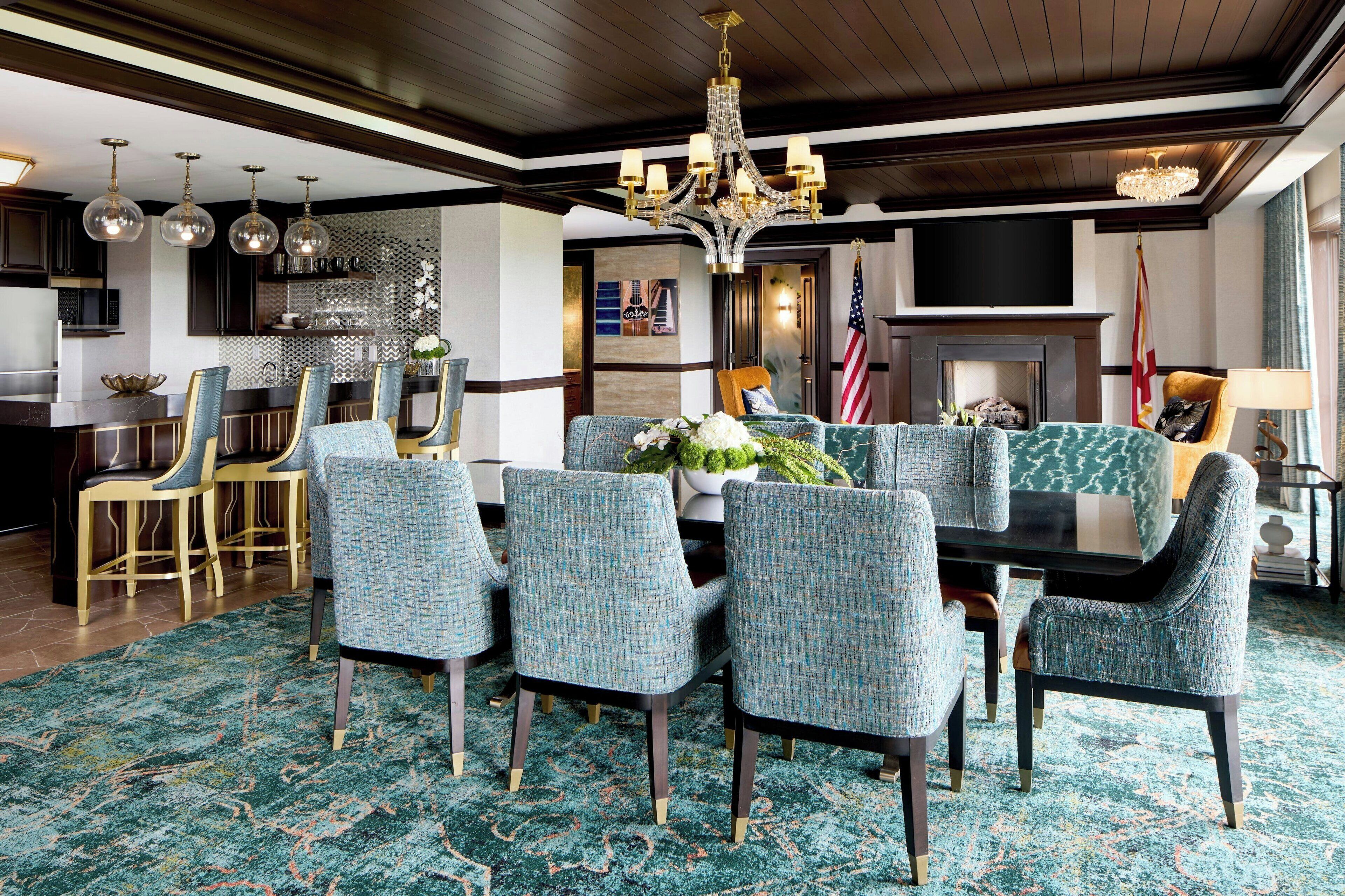 A dining area with a round table, six upholstered chairs, a chandelier, and a patterned carpet.