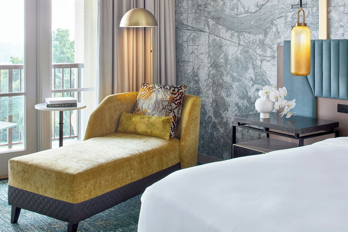 Habitación de hotel con una cama grande, un sofá con un cojín estampado, una mesita con un libro, una lámpara y un mural con un mapa.